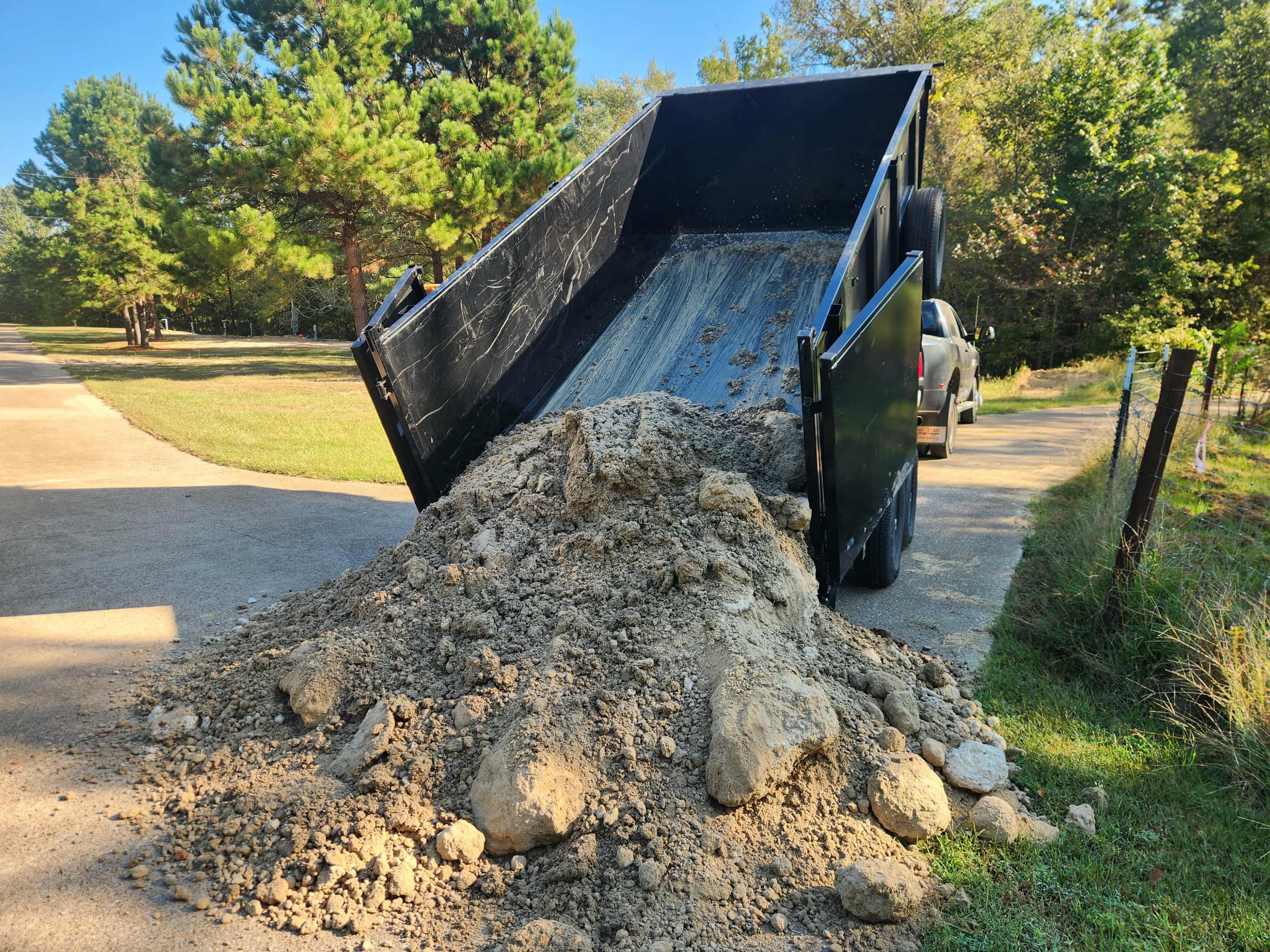 Black dump trailer tipping a large mound of soil onto a paved driveway.